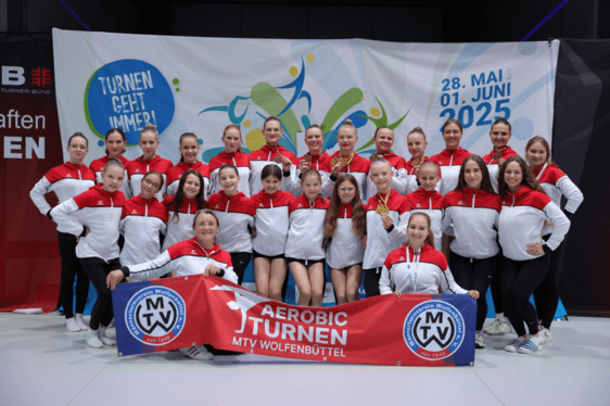 Mannschaftsfoto der Aerobicturner. Sie stehen vor einem großen Banner des Deutschen Turnfestes in Leipzig.